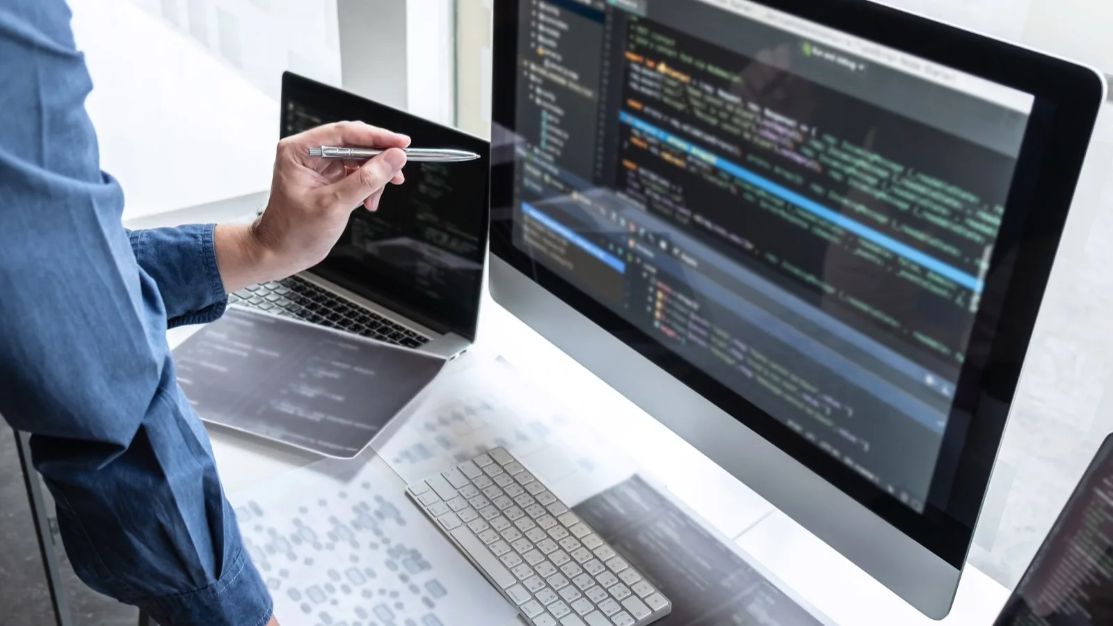Developer pointing at code editor on large monitor next to laptop